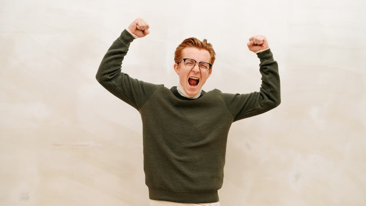 Young man in glasses and sweater jubilantly raises fists indoors, expressing joy and success.